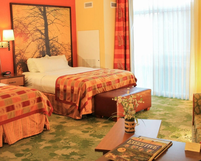 A warmly lit hotel room with two double beds, orange and yellow bedding, large tree artwork above the beds, a green patterned sofa, a TV on a dresser, and a coffee table with a magazine and flowers.