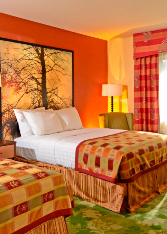 A brightly lit hotel room with two double beds, colorful bedspreads, large tree artwork behind the beds, a desk, lamp, chair, and a window with red patterned curtains.