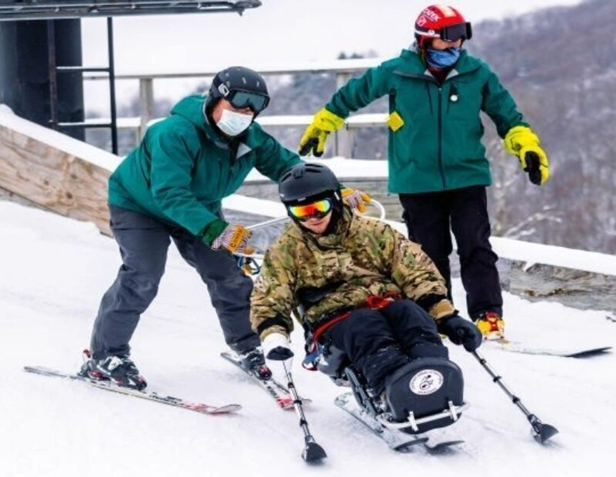 Two people in green jackets assist a person using a sit-ski down a snowy slope. All wear helmets; one helper skis behind, the other stands nearby. Snowy trees and ski lift are visible in the background.