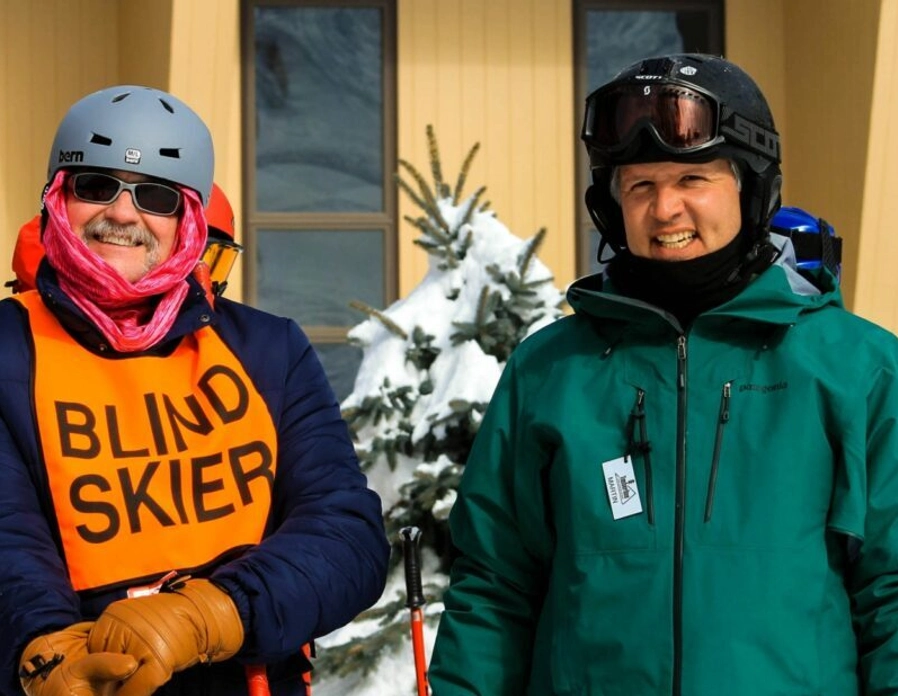 Two people stand outside in ski gear; one wears an orange vest labeled "Blind Skier" and pink scarf, while the other wears a green jacket. Snow and skis are visible in the background. Both are smiling.