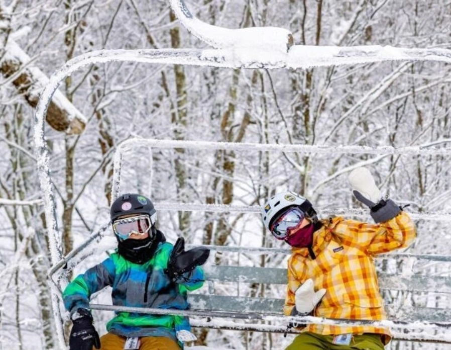 Two snowboarders in colorful winter gear ride a snow-covered ski lift, surrounded by trees coated in fresh snow. One waves at the camera while both appear to enjoy the wintry scenery.