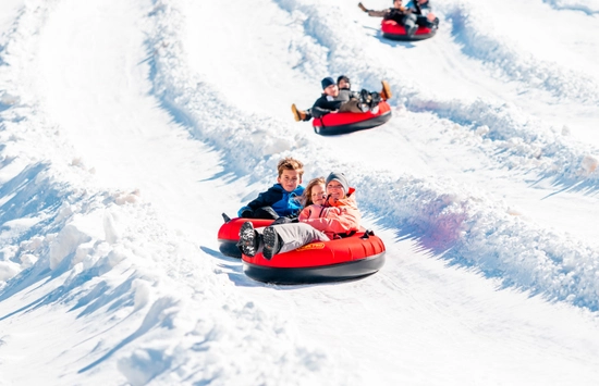 Children in winter coats ride red snow tubes down a snowy hill, smiling and having fun, with several more kids tubing in the background on parallel tracks.