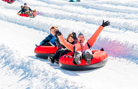 Three people bundled in winter clothes ride red snow tubes down a snowy hill, smiling and waving with excitement. Other people on snow tubes glide down nearby lanes in the background.