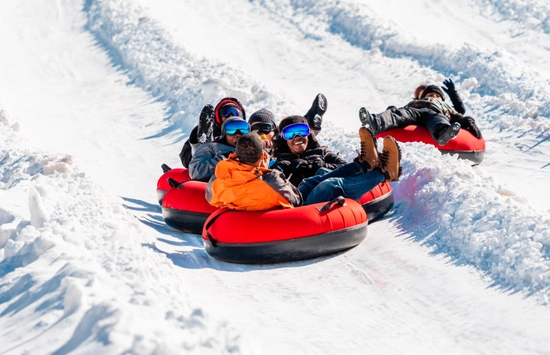 Four people in winter gear slide down a snowy hill in red inflatable snow tubes, smiling and laughing on a sunny day. Snow walls line the track on both sides as they enjoy the outdoor activity.