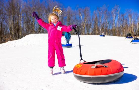 A young child in a bright pink snowsuit smiles and jumps in the snow while holding onto a red snow tube, with a sunny blue sky and leafless trees in the background. Other people are tubing in the distance.