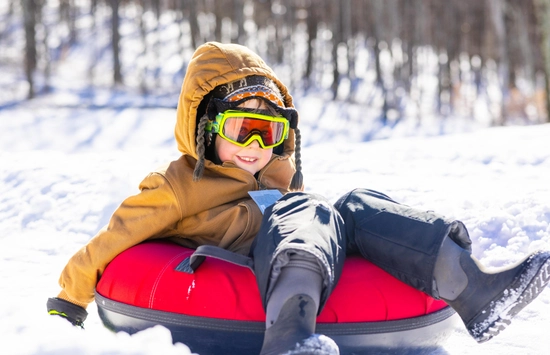 A child wearing a brown hooded jacket, ski goggles, and black boots sits on a red snow tube in a snowy outdoor setting with trees in the background, smiling and enjoying the winter day.