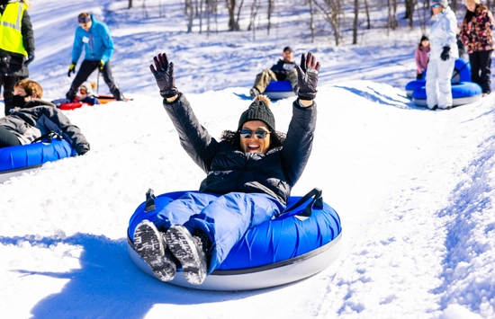 A person wearing winter clothes and sunglasses slides down a snowy hill on a blue snow tube, smiling with arms raised. Other people are tubing and standing in the background.