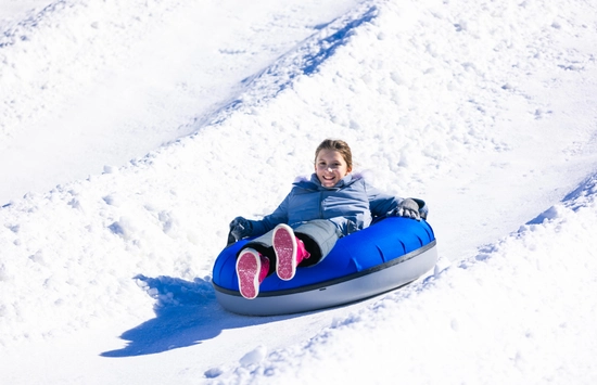 A person wearing a light blue jacket and pink shoes is smiling while sliding down a snowy hill on a blue inflatable snow tube.