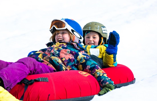 Two children wearing helmets and colorful winter clothes laugh and enjoy riding together on a red snow tube down a snowy hill.